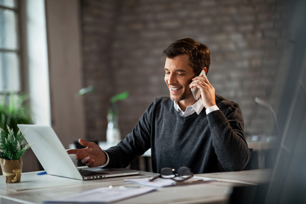 Happy businessman communicating over mobile phone and working on laptop while being at work.